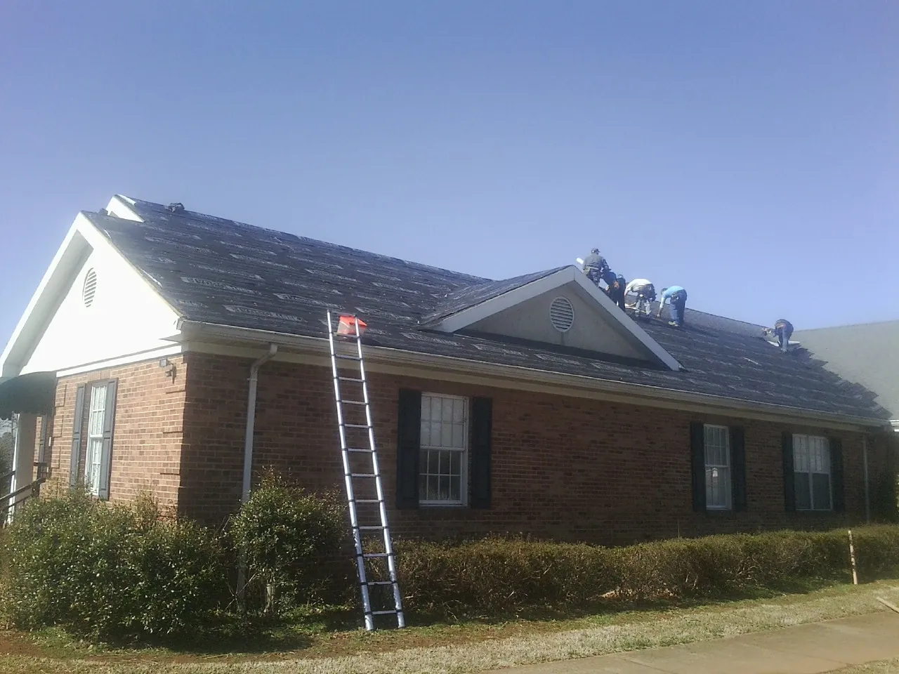 Crew tearing shingles off a brick ranch roof with a ladder set — Emergency Roof Repair work by our crew in Brookhaven